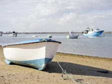 Un bote de pesca sobre la arena en una playa de Huelva y al fondo barquitos pesqueros en la mar.